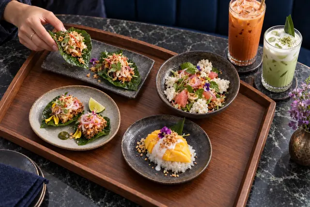 A daytime walnut tray of Thai leaf bites, crisp fish, pomelo salad and mango sticky rice, with a guest lifting a leaf bite.